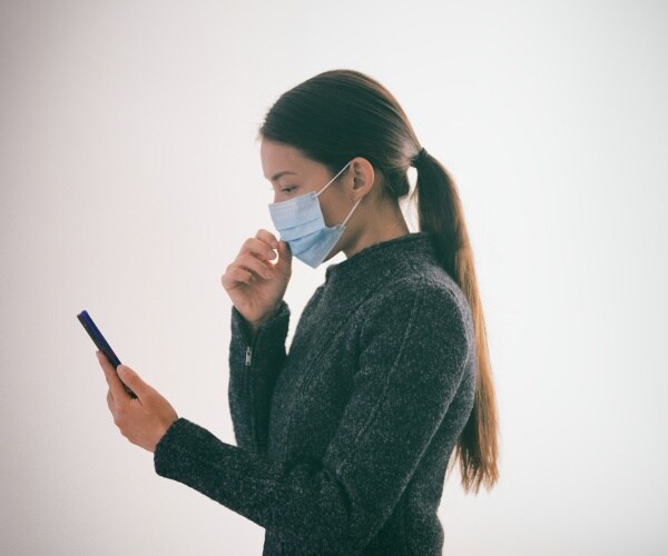 girl wearing a gray jacket and face mask touching her mask and holding up her smart phone