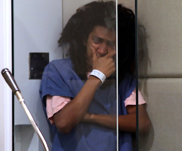 a man with bushy black hair stands with his right hand covering his face in a court room