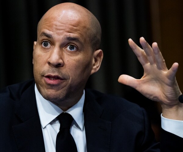 booker in a black suit and tie with one hand raised up
