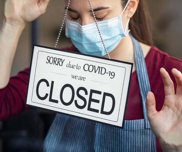 a shopkeeper in a surgical masking looking sad behind a sorry we are closed sign