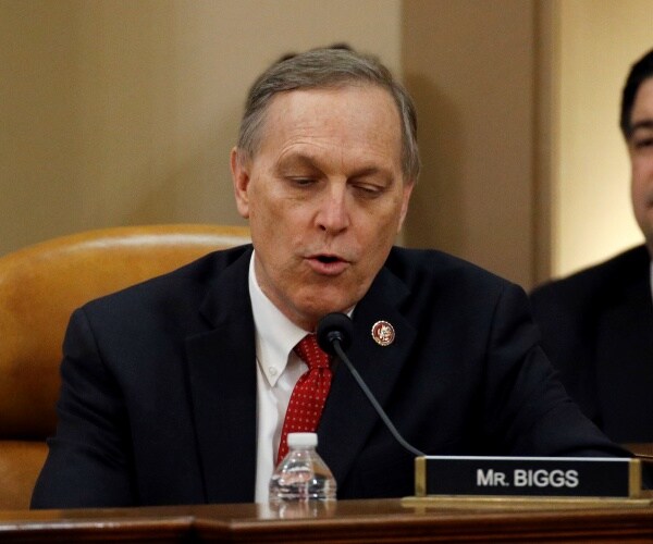biggs in a suit and red tie looking down while speaking at a hearing