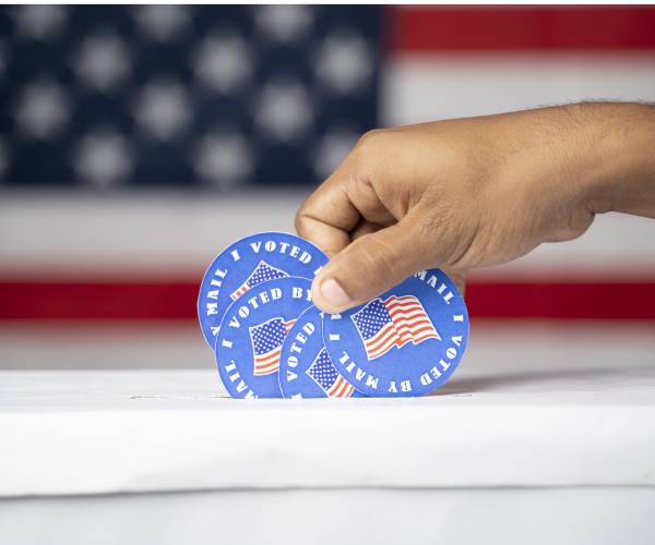 a hand stuffing a ballot box with an american flag in the background