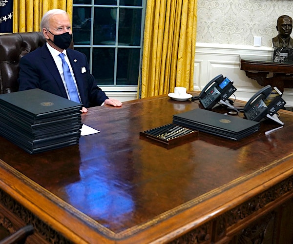 president joe biden sits behind the resolute desk with a stack of executive orders to sign in the oval office