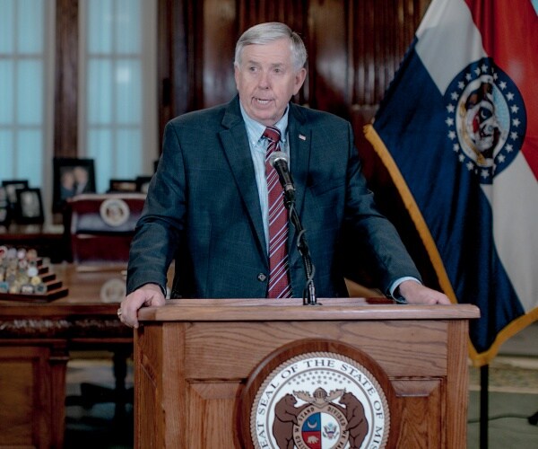 parson at a podium wearing a suit and burgundy striped tie with a missouri state flag behind him