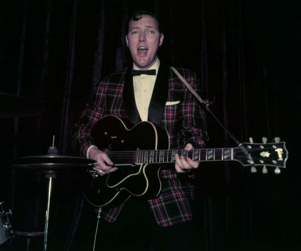 bill haley is shown in a plaid suit jacket with his guitar in london in 1957