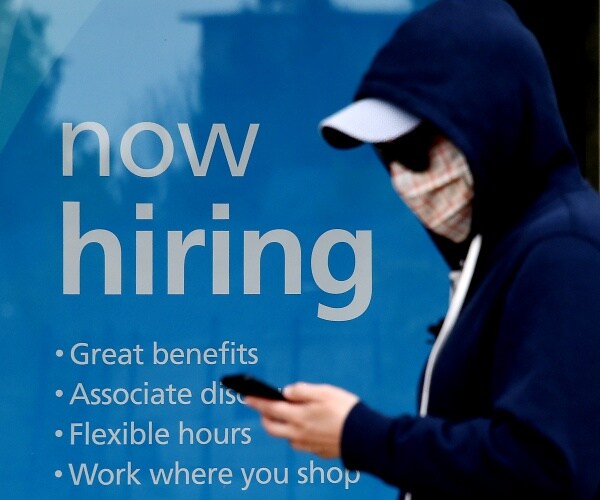 man in a hoodie and mask walks by a hiring sign