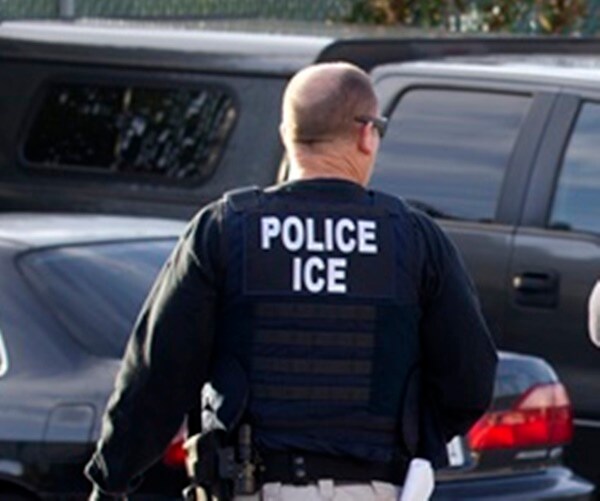 an ice officer in a bulletproof vest walks away in a parking lot