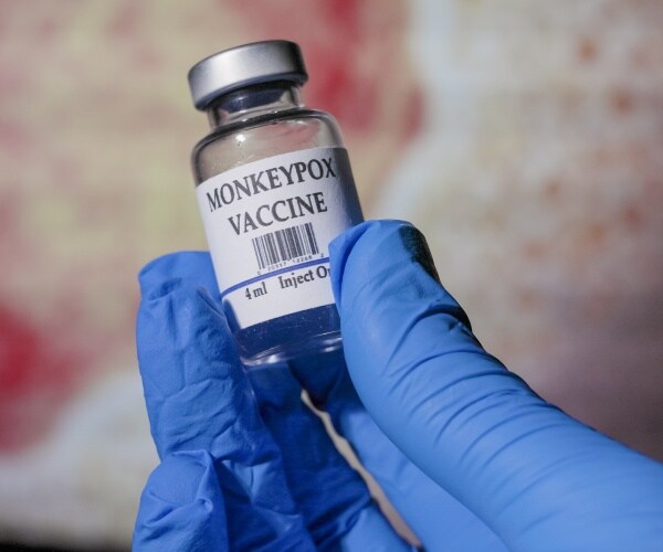 a hand with surgical glove on holding monkeypox vaccine and syringe in front of a picture of monkey pox rash