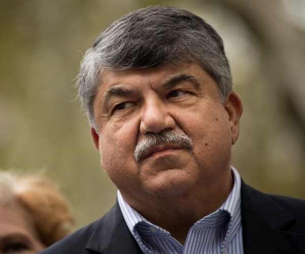 trumka in a suit and blue and white striped shirt standing outside
