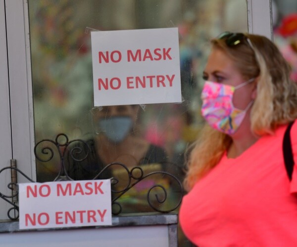 woman in a neon orange top and pink patterned mask walks by a business with a no mask no entry sign