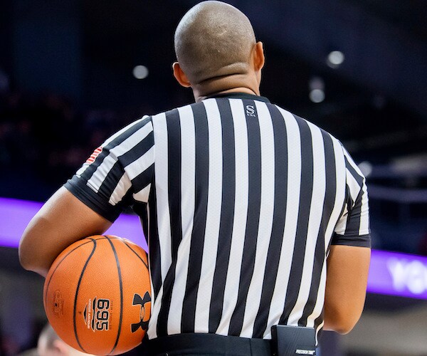 a basketball referee with his back to the camera holds a ball under his left arm as it rests on his left hip