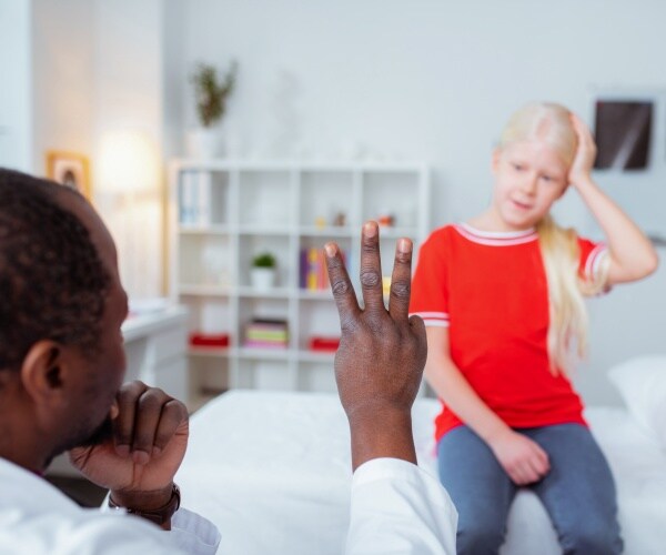 young girl sitting up on exam table holding her head while doctor holds up three fingers to her to look at