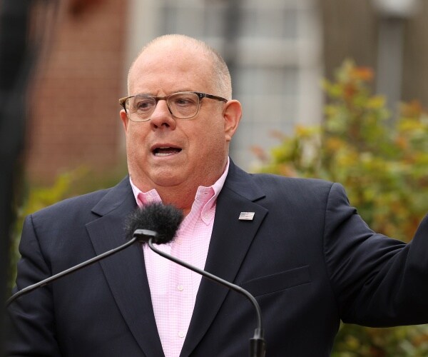 hogan in a black suit and pink dress shirt speaking outside