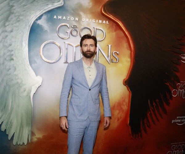 david tennant, standing in front of a poster background of the amazon prime production "good omens"