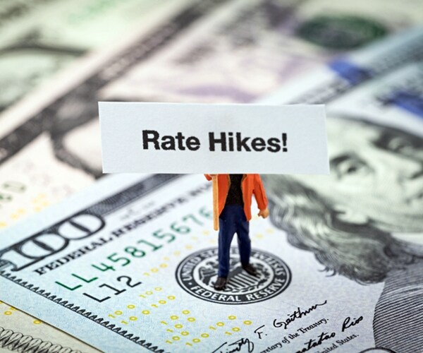 miniature man holding Rate Hikes sign standing on US Federal Reserve emblem on dollars banknote.