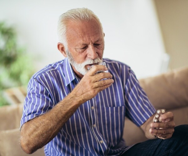 man sitting on couch, holding a glass of water in one hand, putting pill in mouth with other