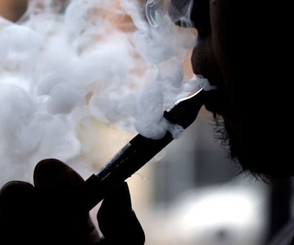 a man puffs vape smoke out of an e-cigarette in the foreground shadow