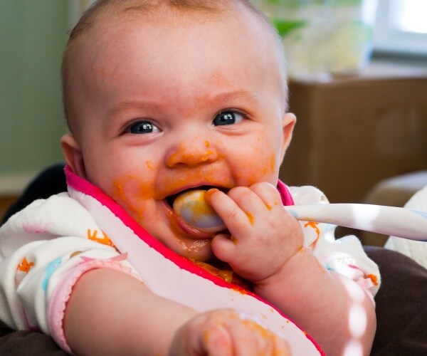 baby with baby food on face and spoon in mouth smiling at camera