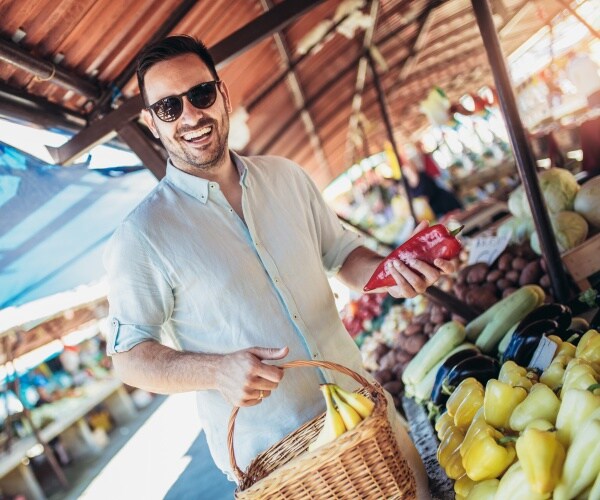 man smiling as he picks up vegetables at an open air market