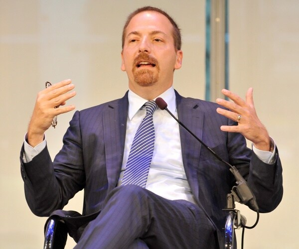 chuck todd speaking onstage wearing a suit and striped tie with his hands raised