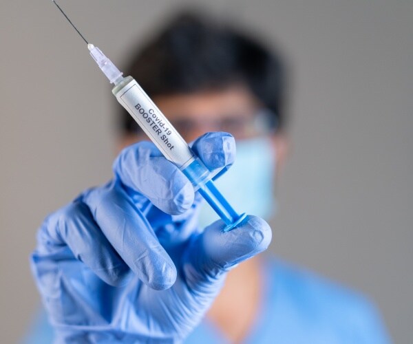 a healthcare worker in scrubs and mask holding up a syringe to the camera, labeled 'covid booster'