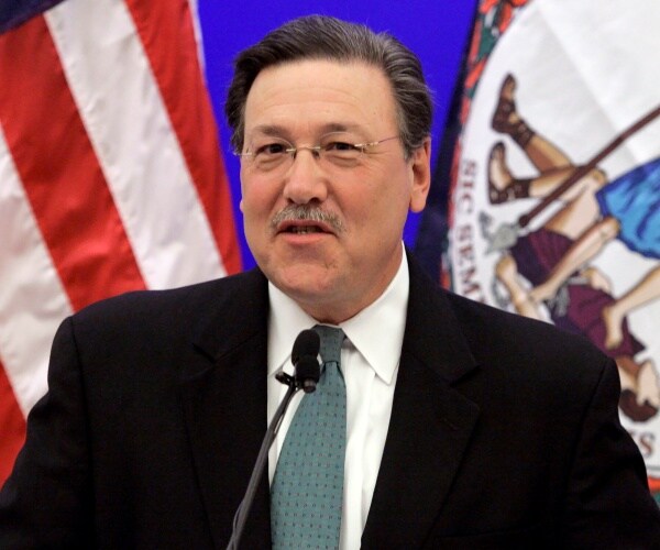 domenech in a black suit and teal patterned tie speaking in front of an american flag