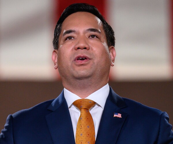 sean reyes speaks at a podium wearing a blue suit and an orange tie