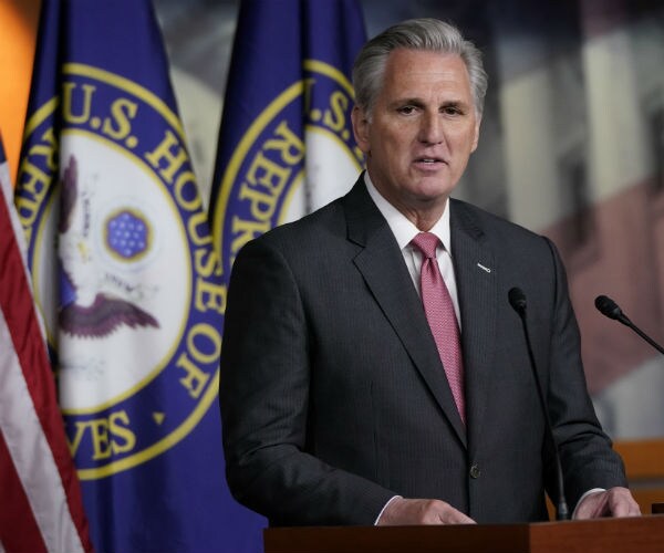 kevin mccarthy is seen in a dark suit, white dress shirt and red tie while speaking to the press