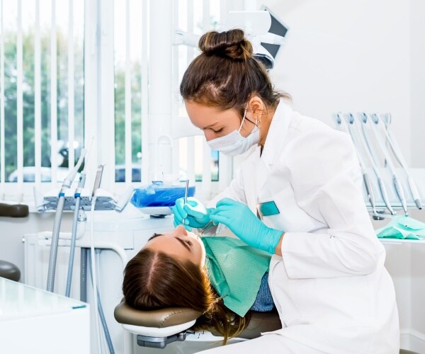 dentist in a white coat and face mask looks at a woman's teeth