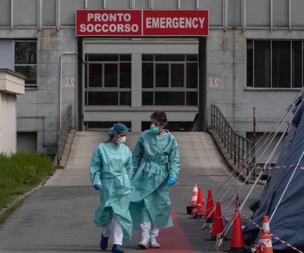 two nurses in gowns and masks outside a hospital in italy reading emergency and pronto soccorso