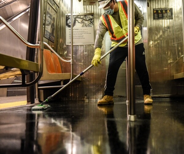 worker in a yellow jacket and mask mops the floor of a subway car in nyc