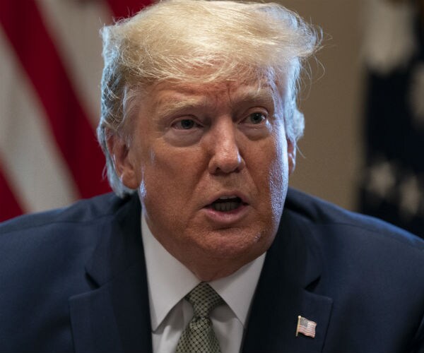 donald trump is shown speaking in a blue suit, dress shirt and tie with the us flag on his lapel