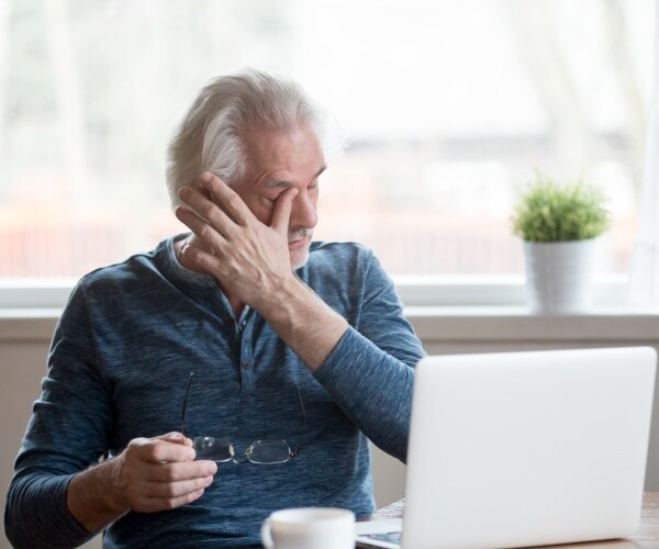 man in front of his computer, with eyeglasses in his hand, rubbing his eyes